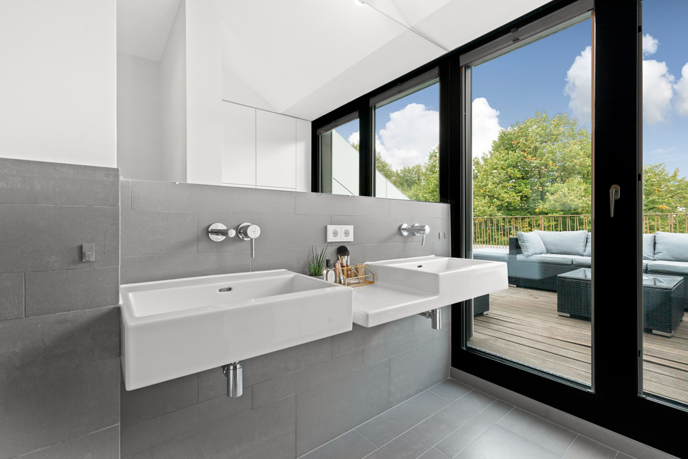 Modernes Bad mit zwei Waschbecken und großen Fenstern, Blick auf eine Terrasse mit Sitzmöbeln.