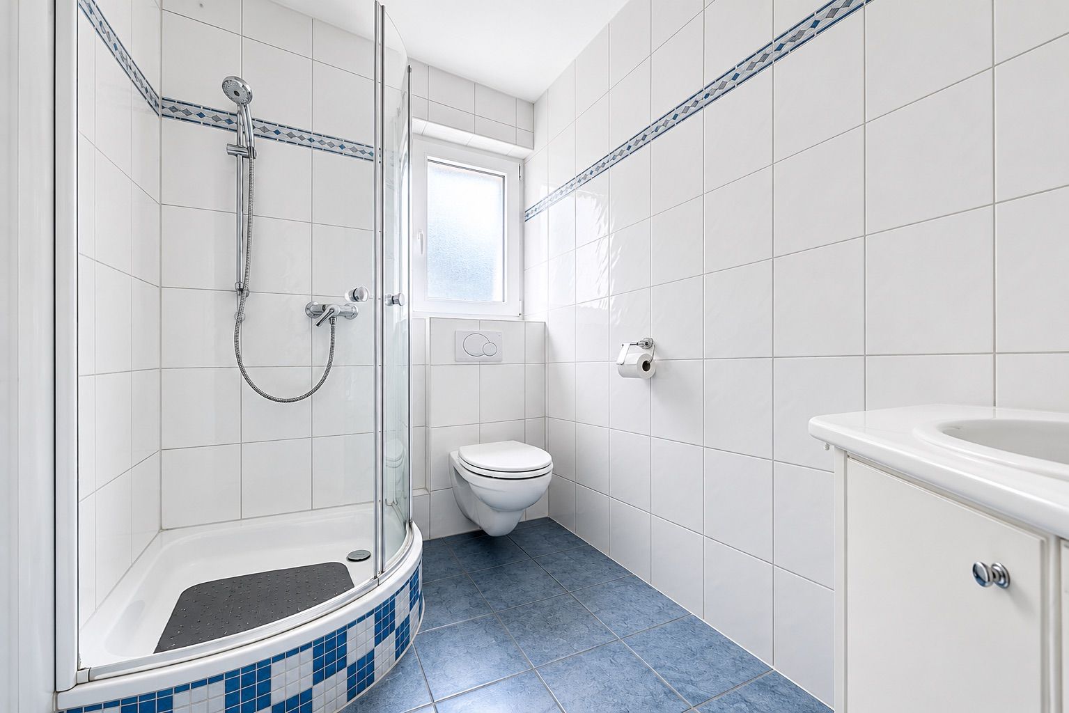 Helles Bad mit Eckdusche, WC, Waschbecken und Fenster, blaue Fliesen am Boden.