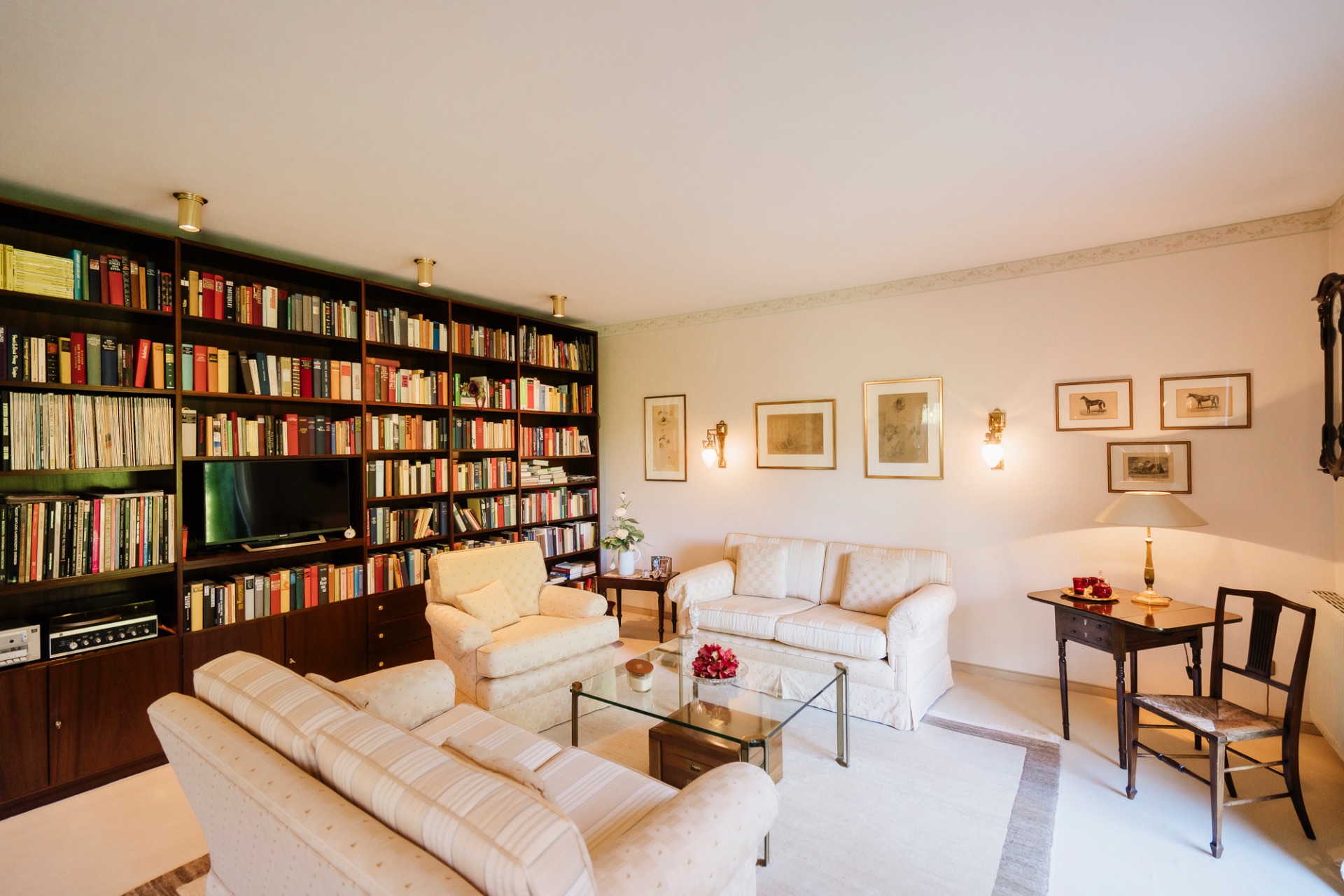 Helles Wohnzimmer mit Bücherwand, beigen Sofas, Glastisch und Schreibtisch mit Stuhl.