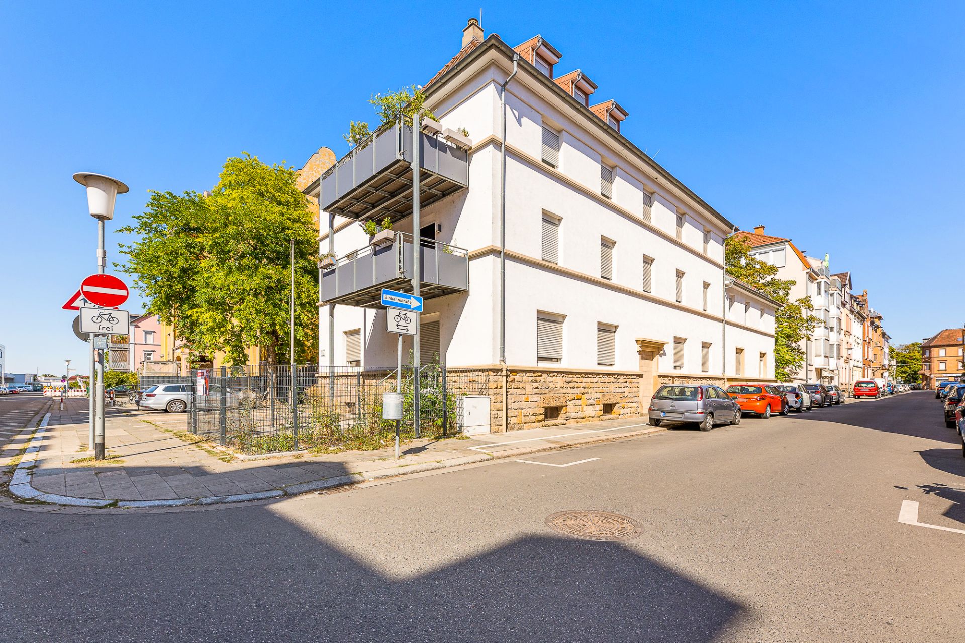 Eckhaus mit weißen Fassaden, mehreren Balkonen und geparkten Autos an einer Straßenkreuzung.
