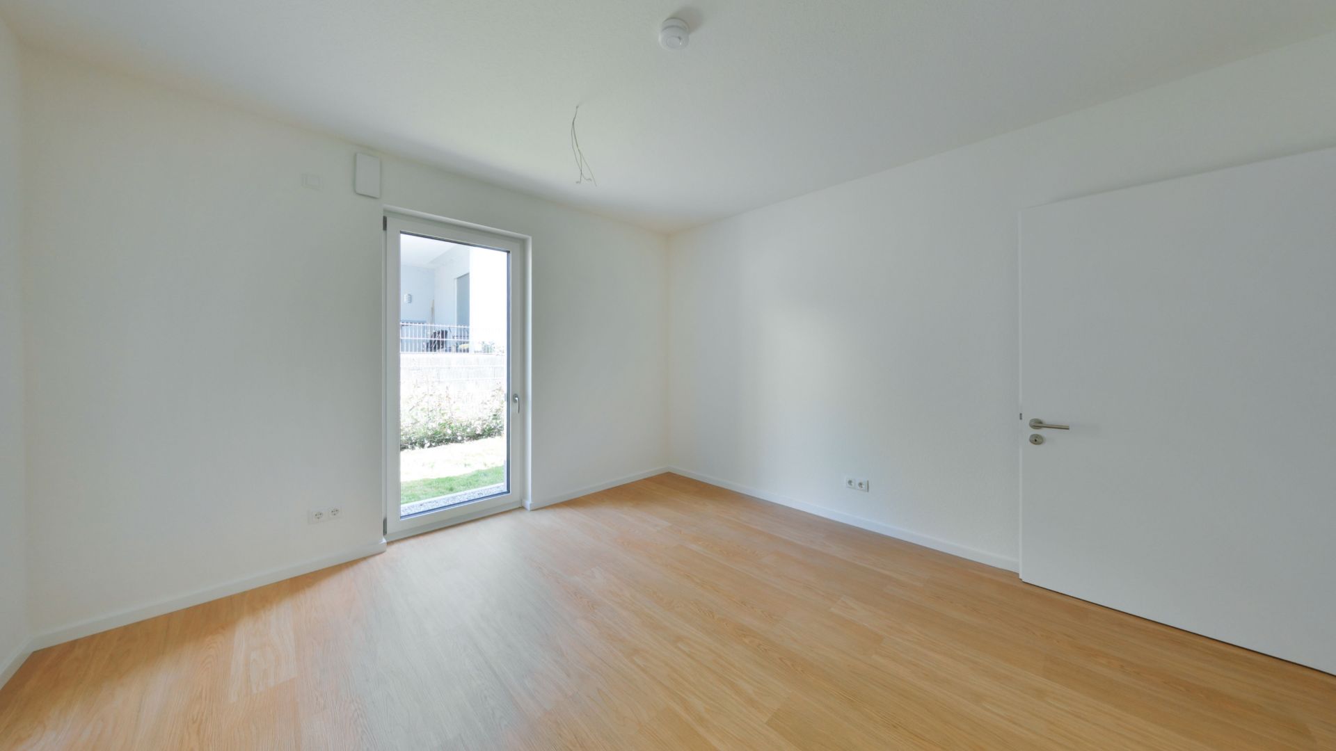 Leerer Raum mit Holzboden, weißer Wand, Tür und Fenstertür zum Garten.