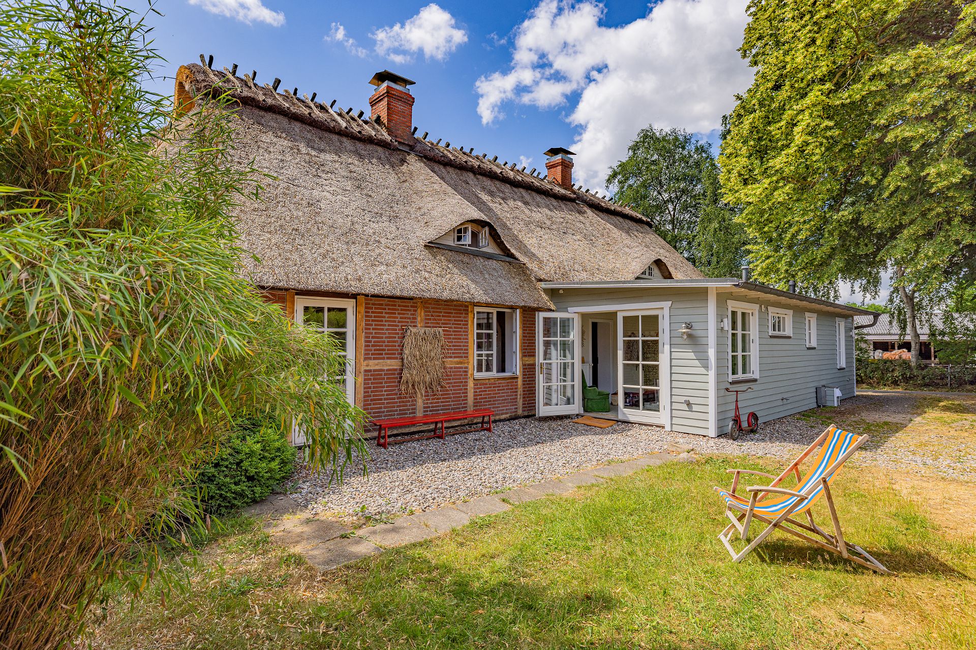 Haus mit Reetdach, roter Bank, Terrasse und Liegestuhl im Garten bei sonnigem Wetter.