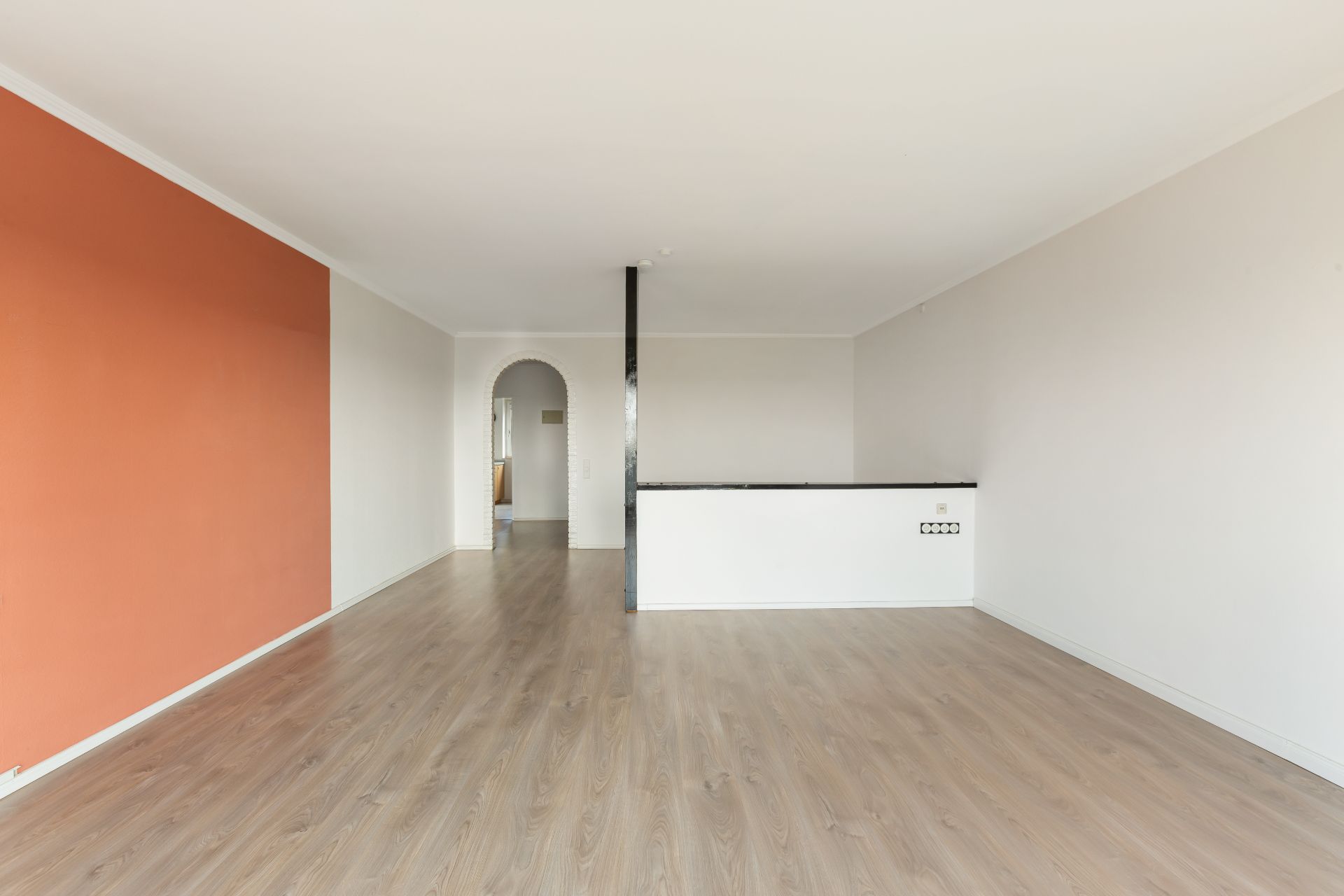 Leerer Raum mit Holzboden, weißer Wand und orangefarbener Akzentwand, offener Durchgang mit Bogen.