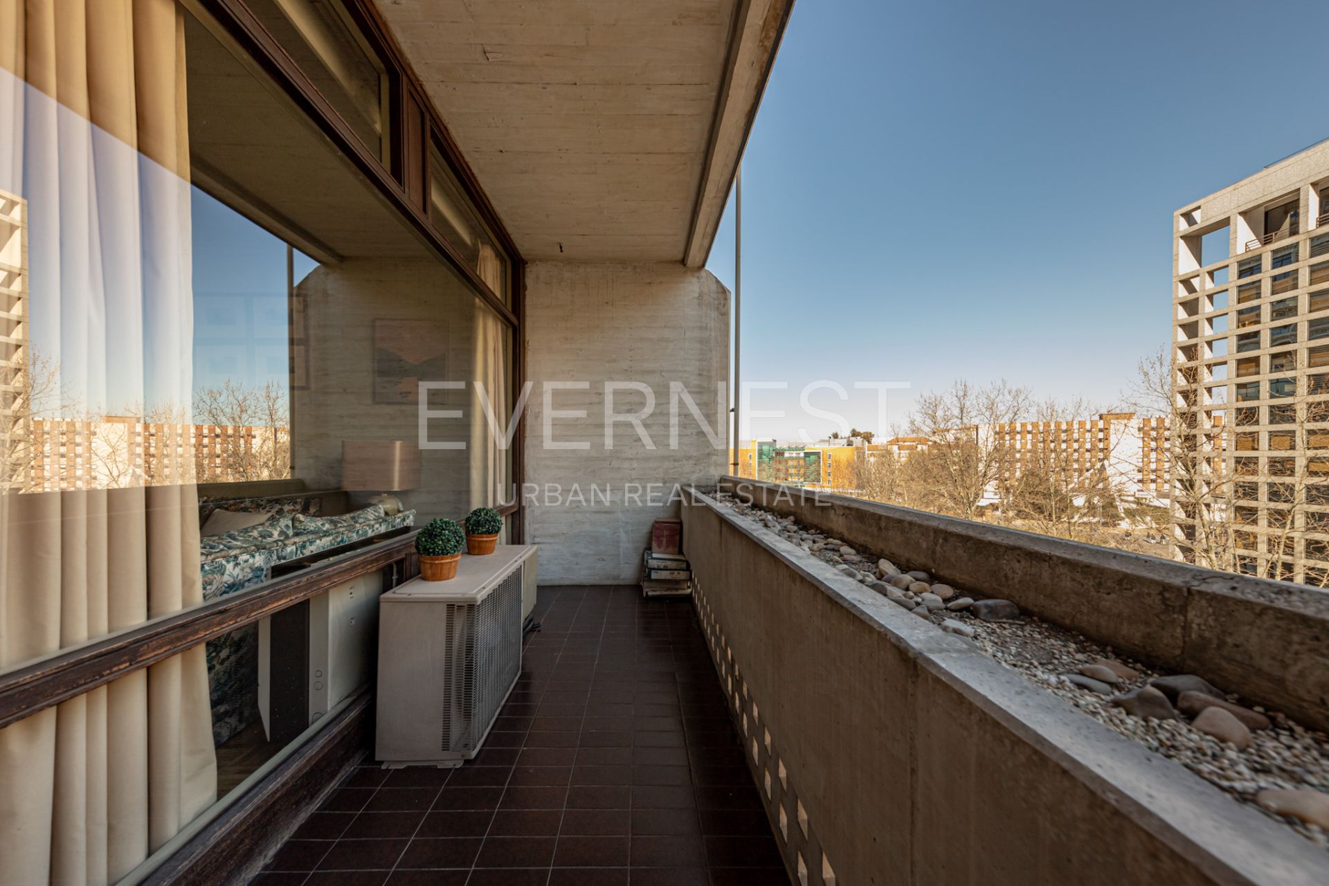 Balkon mit braunen Fliesen, zwei Pflanzen, Steinen am Geländer und Blick auf Wohngebäude.