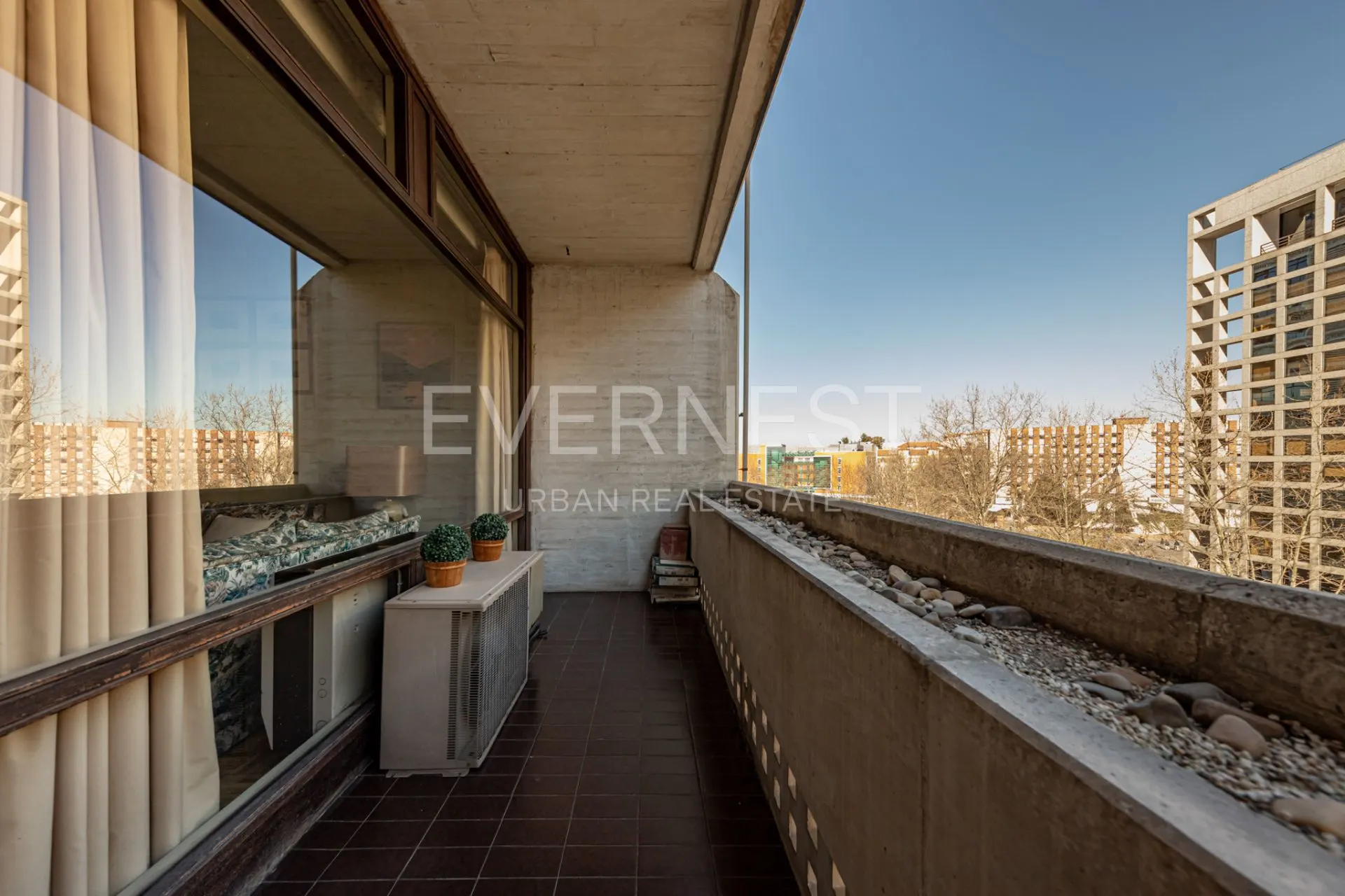 Balkon mit braunen Fliesen, zwei Pflanzen, Steinen am Geländer und Blick auf Wohngebäude.