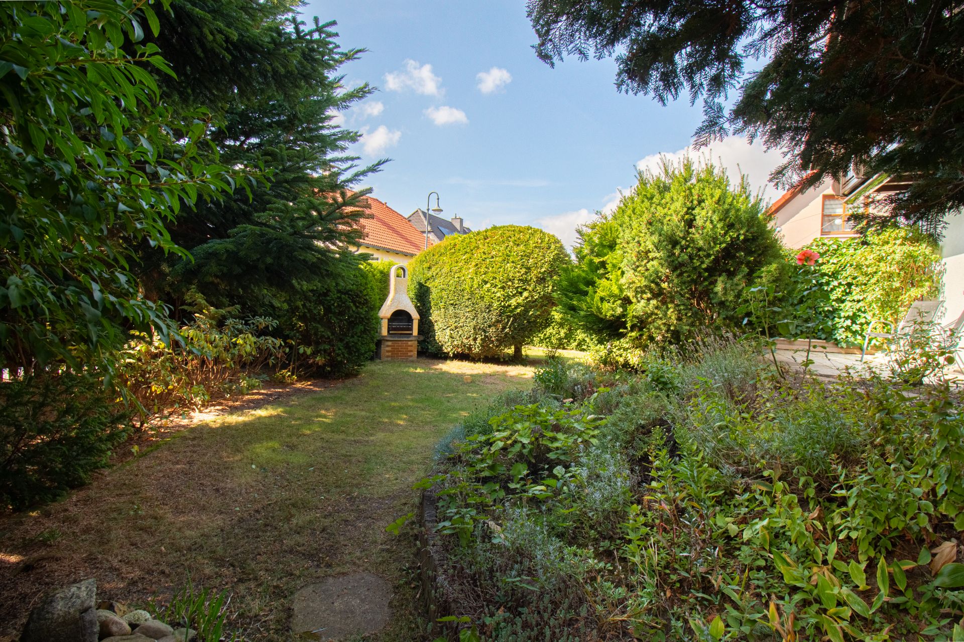 Garten mit Rasen, Bäumen, Sträuchern und einem gemauerten Grill im Hintergrund.