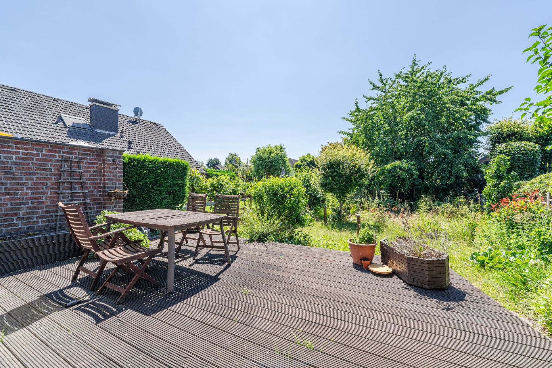 Holzterrasse mit Tisch und Stühlen, umgeben von grünem Garten und Pflanzen.