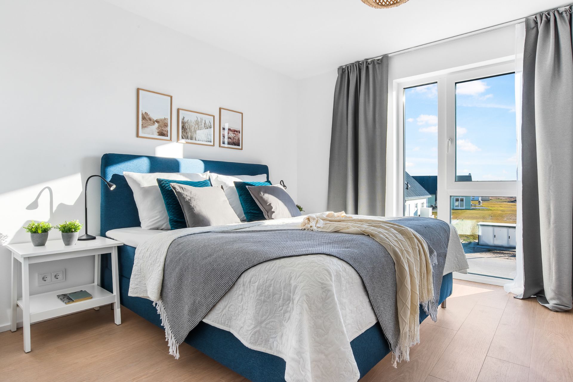 Helles Schlafzimmer mit Doppelbett, Nachttisch, Bildern an der Wand und Fenster mit grauen Vorhängen.