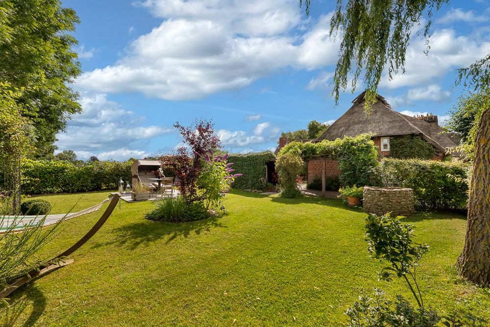 Grüner Garten mit Sonnenliege, Terrasse und Haus mit Reetdach im Hintergrund.