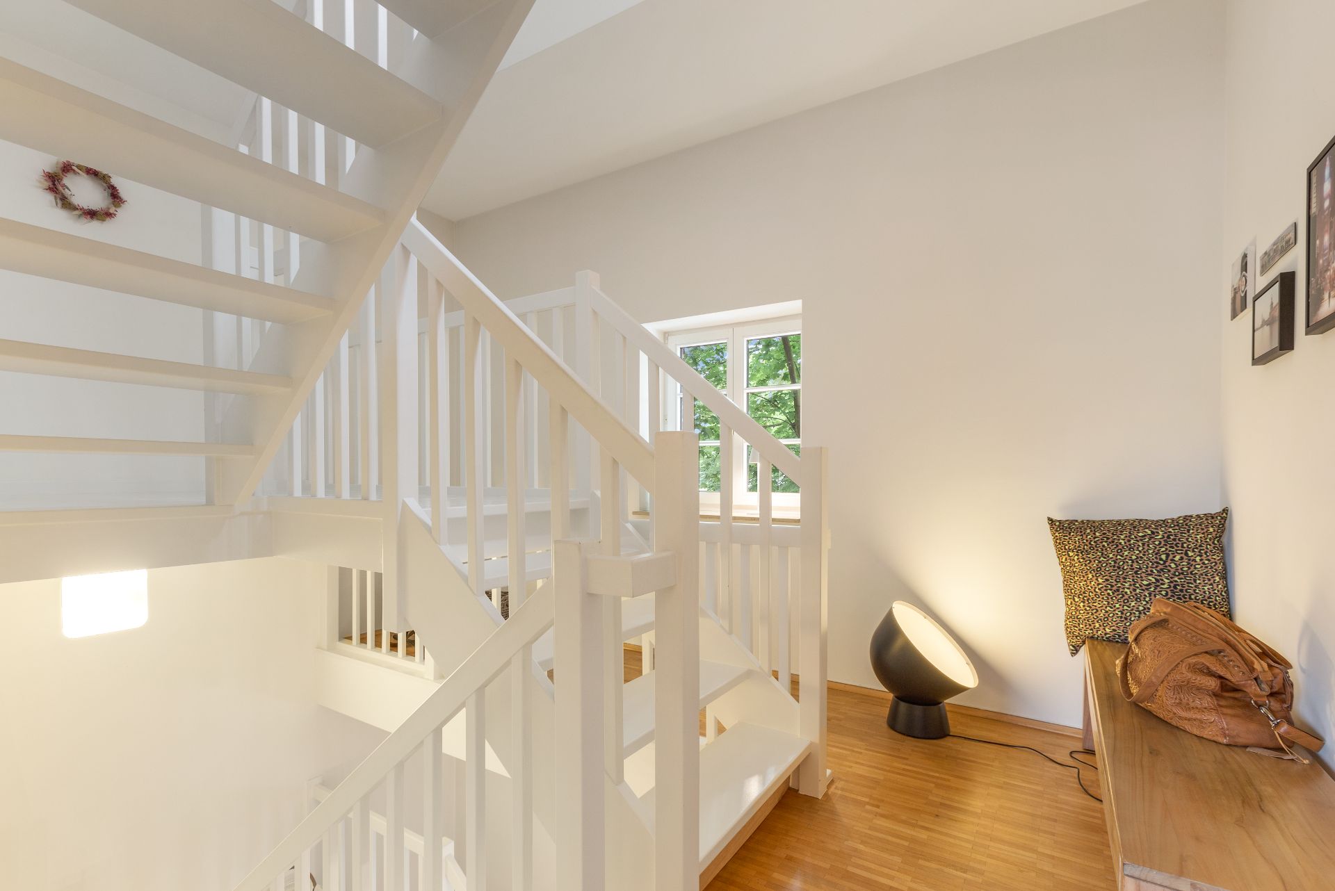 Heller Flur mit weißer Treppe, Holzbank, Kissen, Lampe und kleinem Fenster.