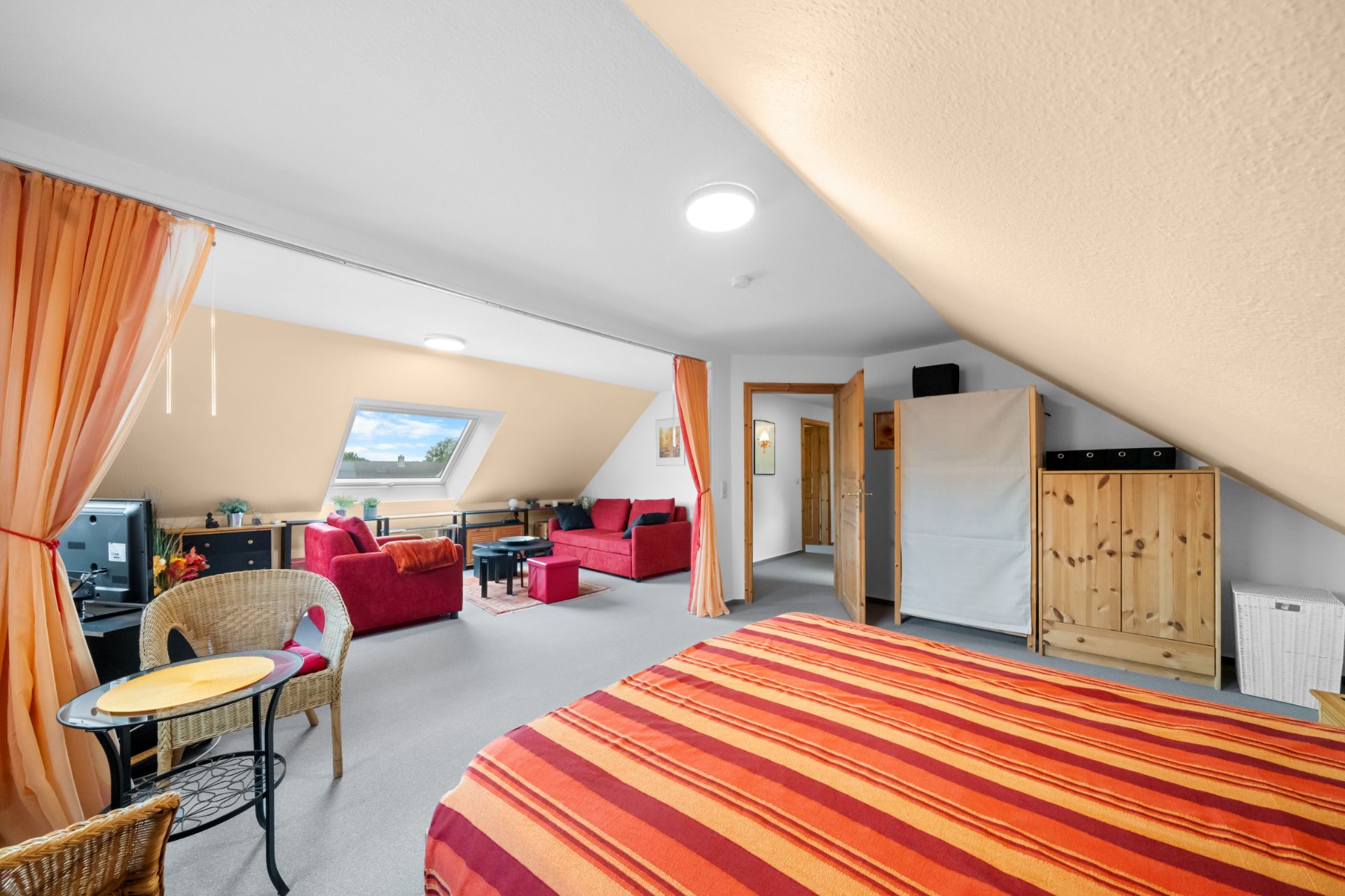 Helles Dachzimmer mit Bett, Sitzbereich, Schreibtisch und Dachfenster, orange Vorhänge trennen Bereiche.