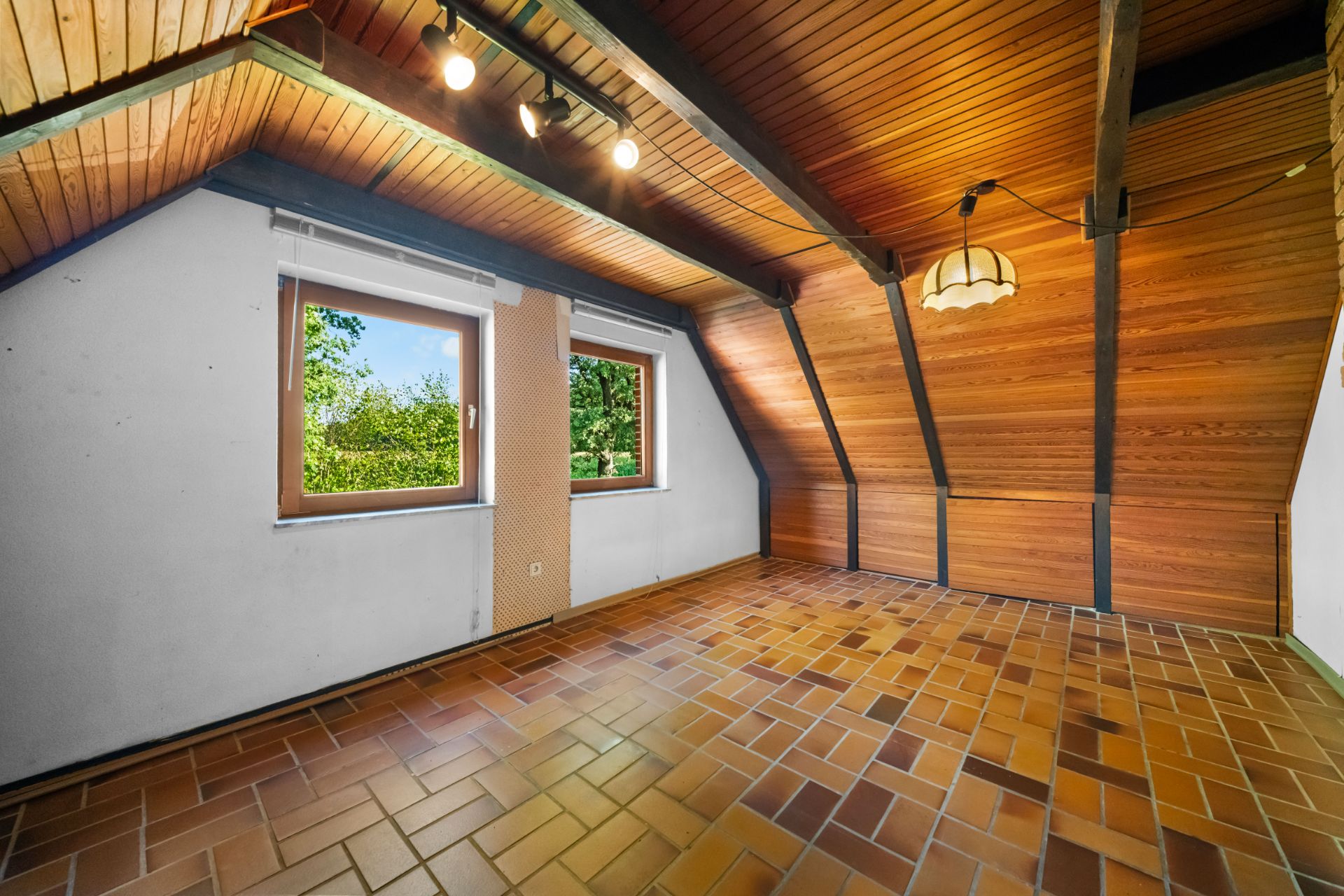Dachzimmer mit Holzdecke, braunem Fliesenboden und zwei Fenstern mit Blick ins Grüne.
