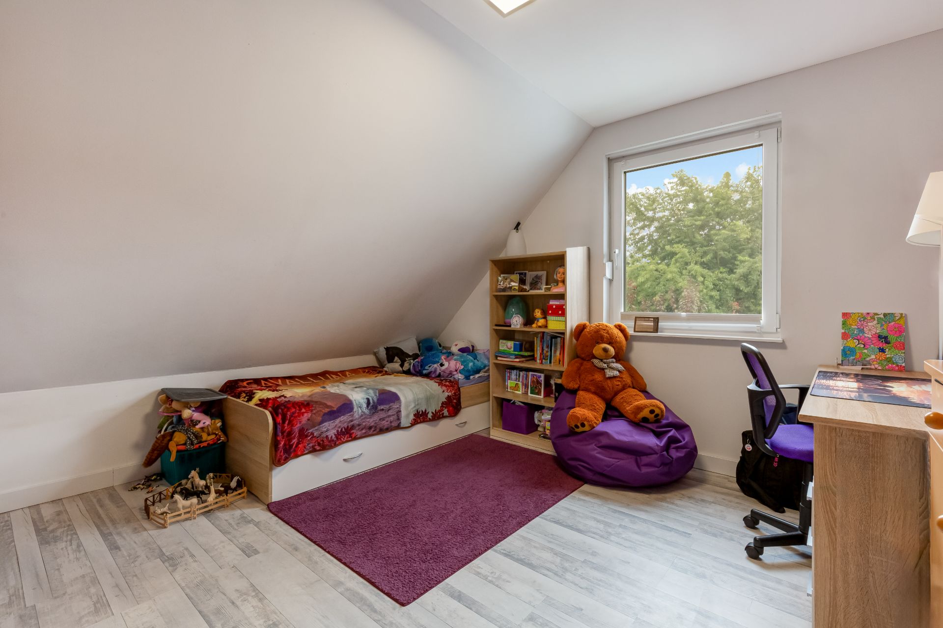 Kinderzimmer mit Einzelbett, lila Teppich, Regal, großem Teddybär und Schreibtisch am Fenster.