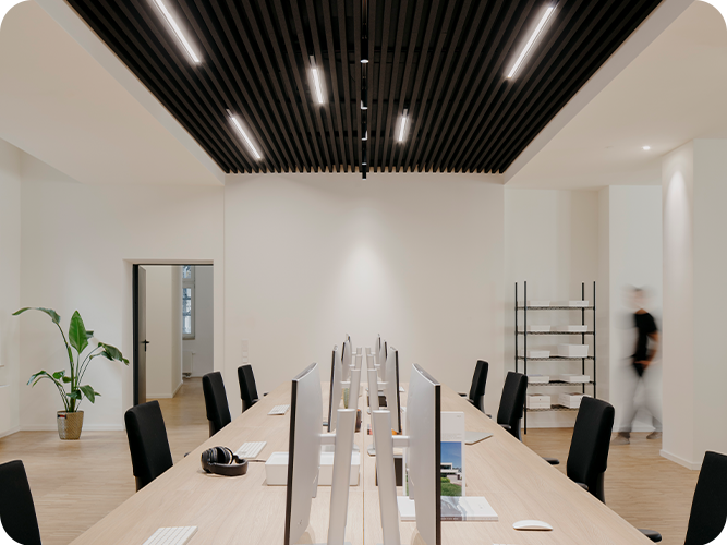 Großer Raum mit langem Schreibtisch, vielen Monitoren, schwarzen Stühlen und Pflanze in der Ecke.