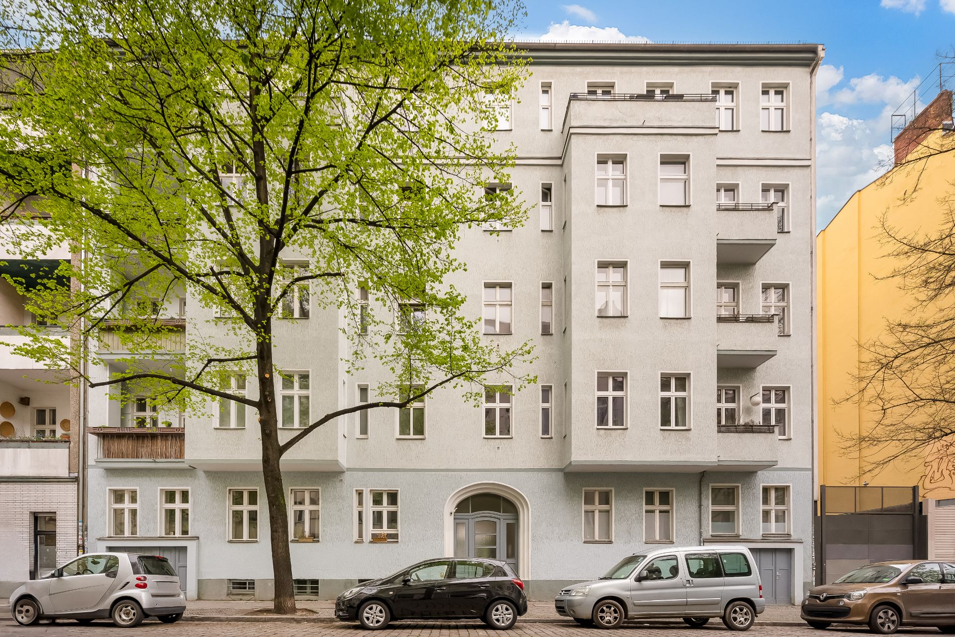 Mehrstöckiges Wohnhaus mit Balkonen, davor Bäume und geparkte Autos an der Straße.
