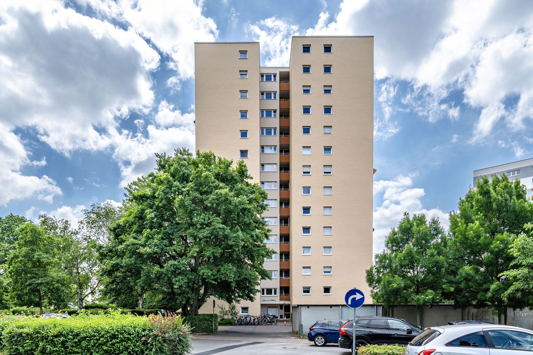 Hohes, helles Wohnhaus mit vielen Fenstern, umgeben von Bäumen und geparkten Autos.