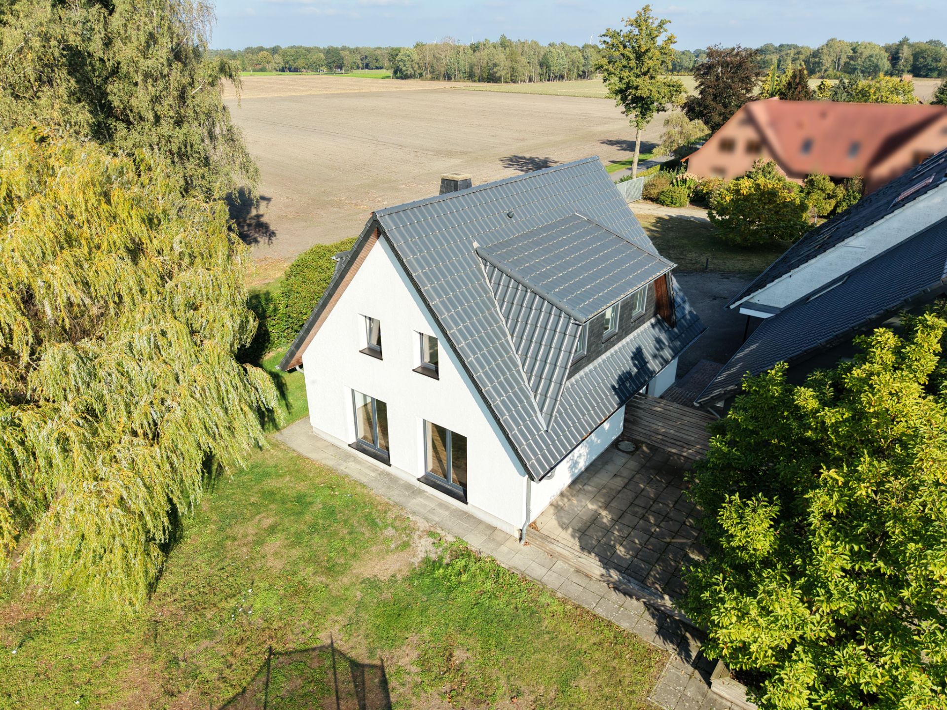 Einfamilienhaus mit dunklem Dach in ländlicher Umgebung, umgeben von Bäumen und Feldern.