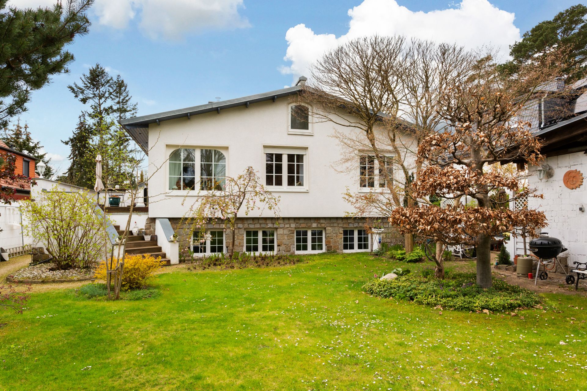 Weißes Wohnhaus mit großem Garten, Bäumen und Terrasse, aufgenommen bei Tageslicht.