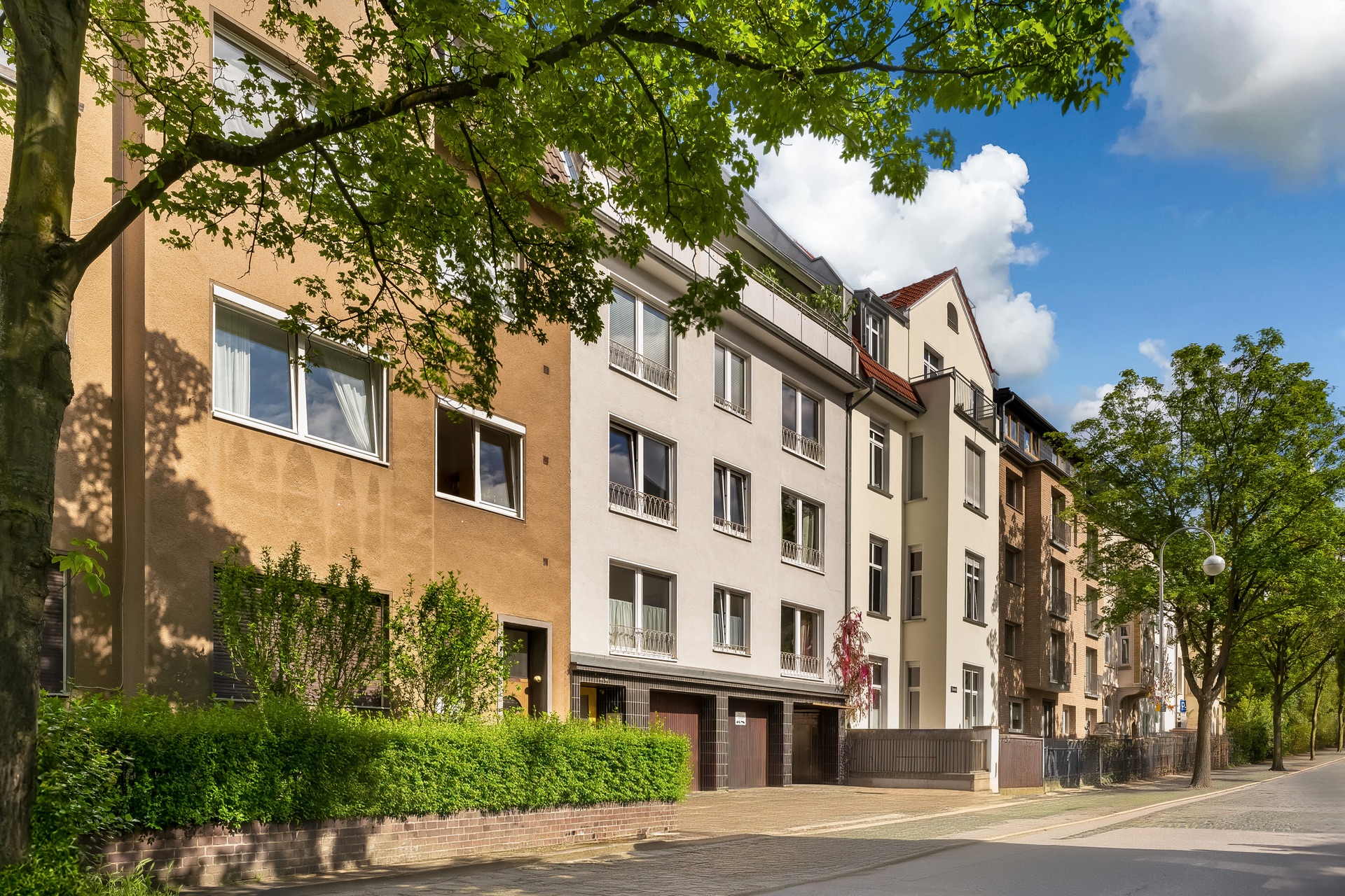 Mehrstöckige Wohnhäuser mit Balkonen an einer ruhigen, von Bäumen gesäumten Straße.