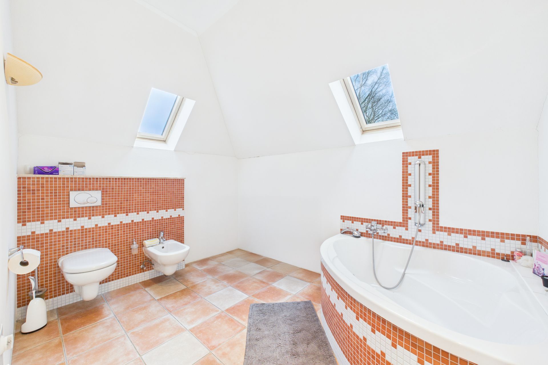 Helles Badezimmer mit Eckbadewanne, Toilette, Bidet, zwei Dachfenstern und orange-weißen Fliesen.