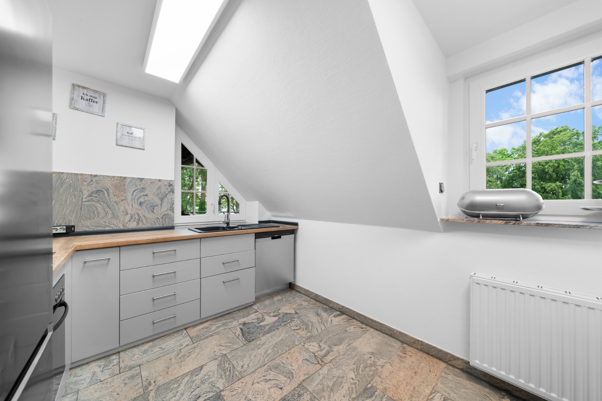 Helle Küche mit schräger Decke, großem Fenster und grauen Schränken.