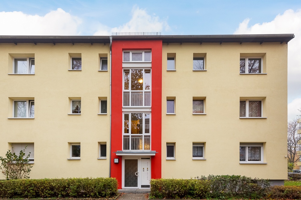 Dreistöckiges Wohngebäude mit gelber Fassade und rotem Hauseingang, viele Fenster sichtbar.