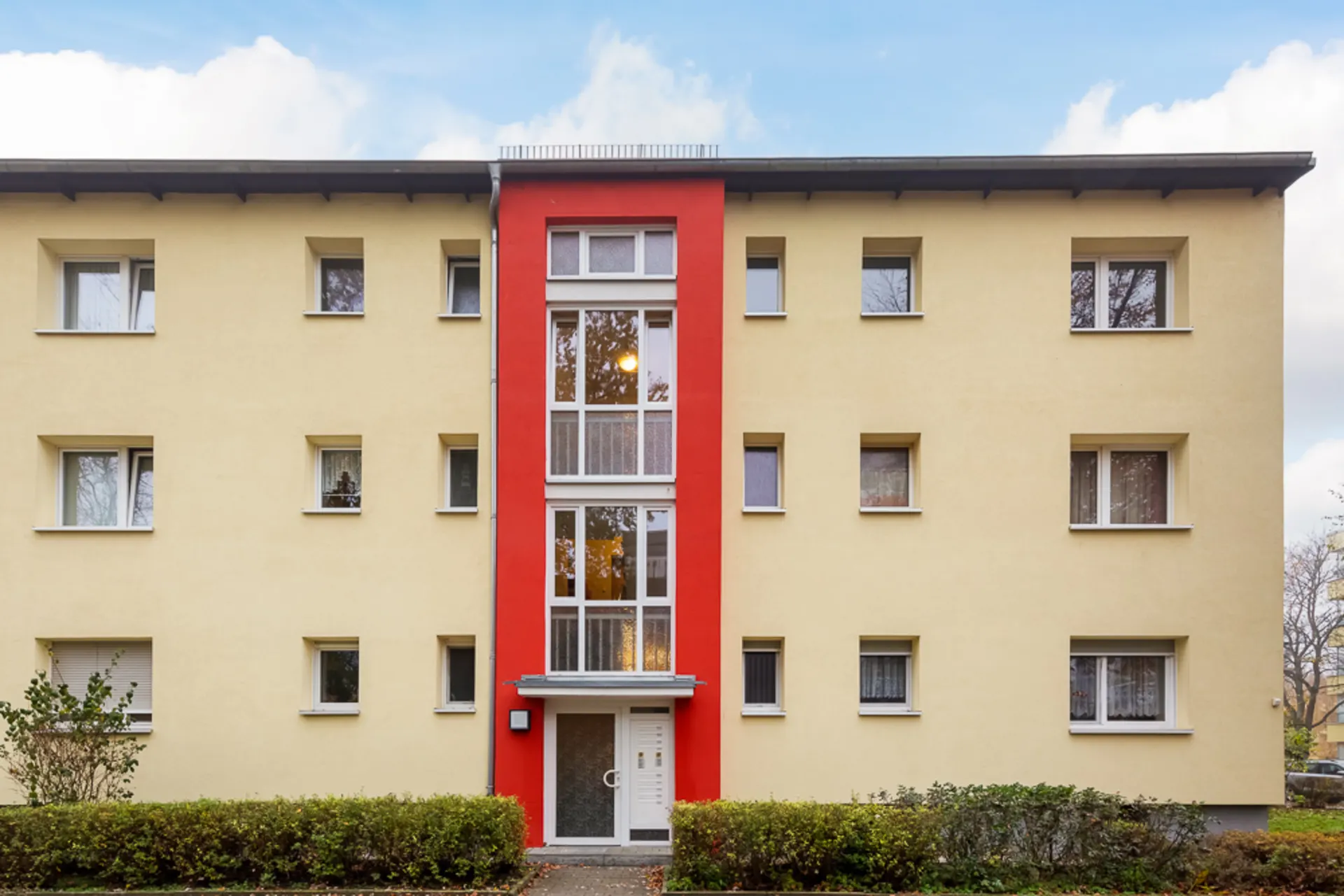 Dreistöckiges Wohngebäude mit gelber Fassade und rotem Hauseingang, viele Fenster sichtbar.