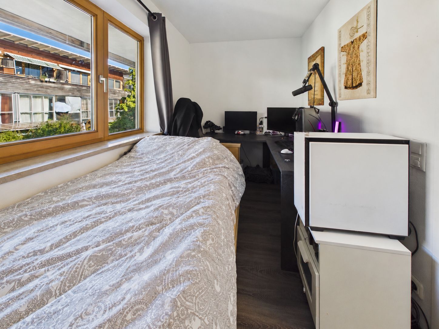 Kleines Zimmer mit Bett, Schreibtisch, Monitoren und großem Fenster mit Blick nach draußen.