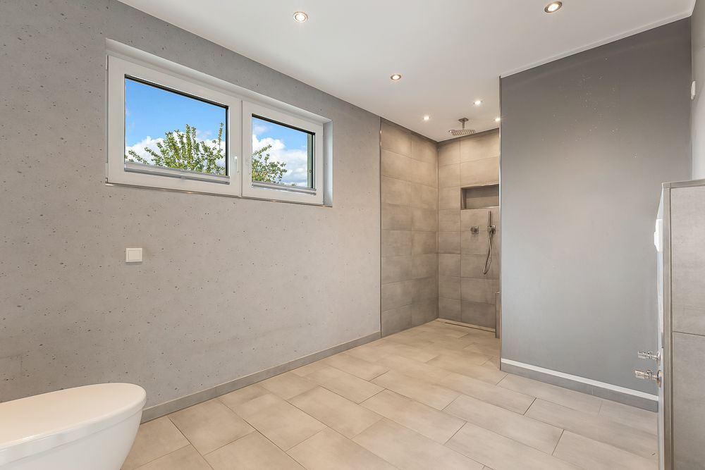 Modernes Badezimmer mit großer, offener Dusche und zwei Fenstern, graue Wand- und Bodenfliesen.