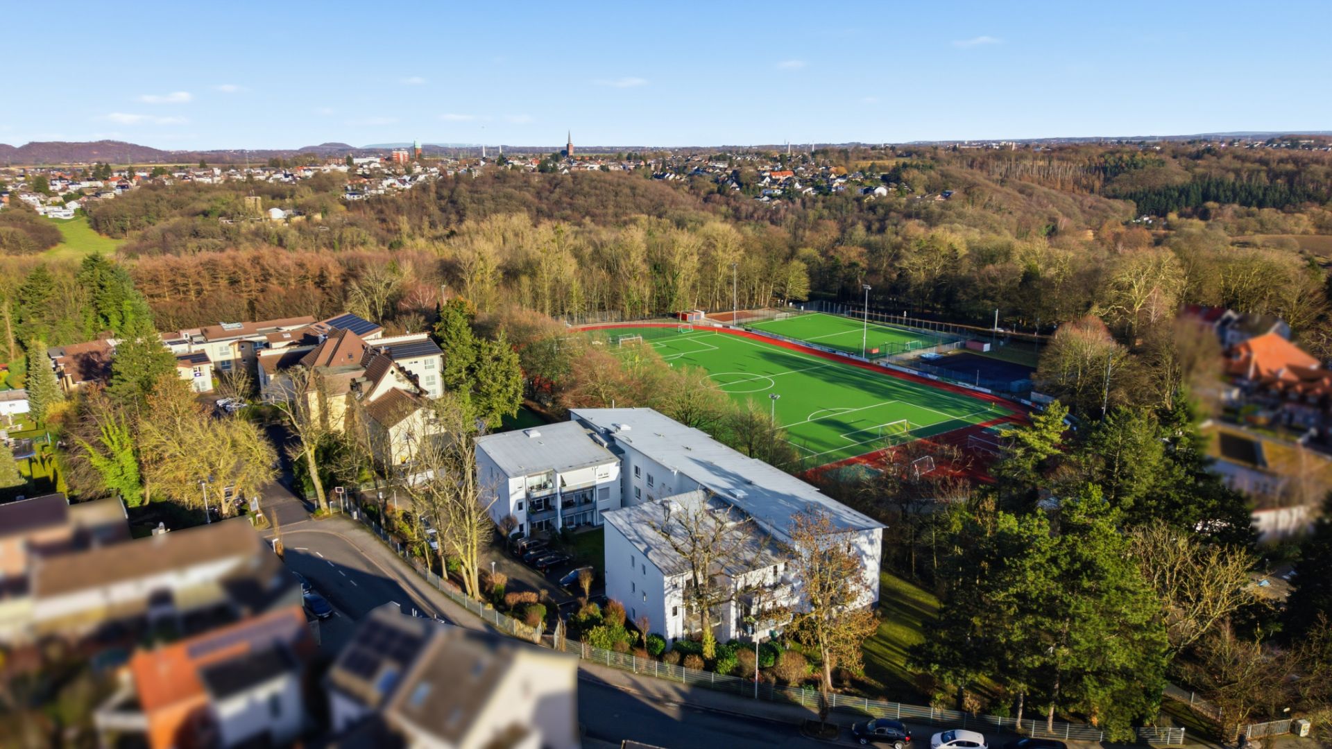 Luftaufnahme von Wohngebäuden neben einem großen, grünen Sportplatz mit umliegenden Bäumen.