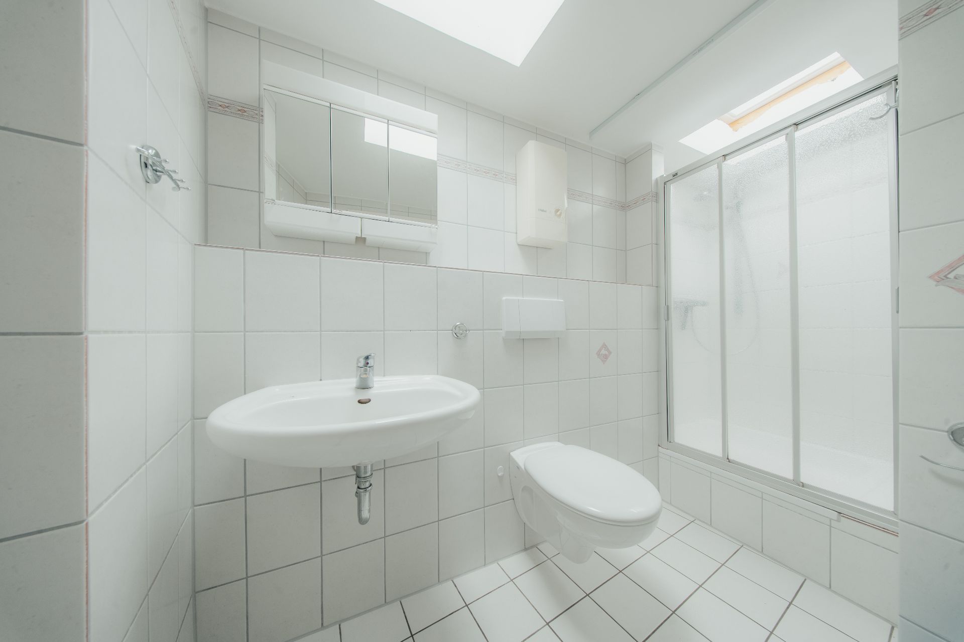 Helles Badezimmer mit Waschbecken, Wandtoilette, Spiegel und Dusche mit Glasabtrennung.
