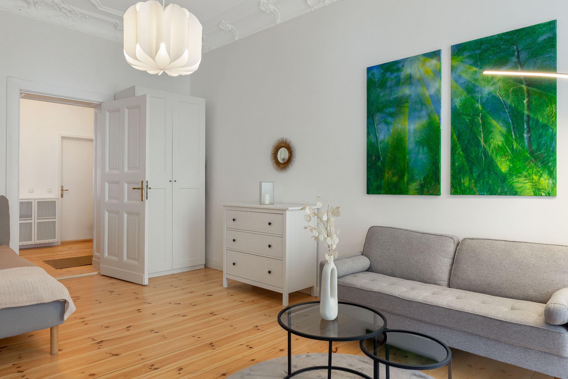 Wohnzimmer mit grauem Sofa, weißen Möbeln, Holzboden und zwei grünen Bildern an der Wand.