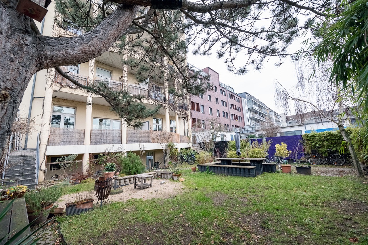 Garten mit Bäumen, Bänken und Pflanzkübeln vor einem Wohnhaus mit Balkonen.