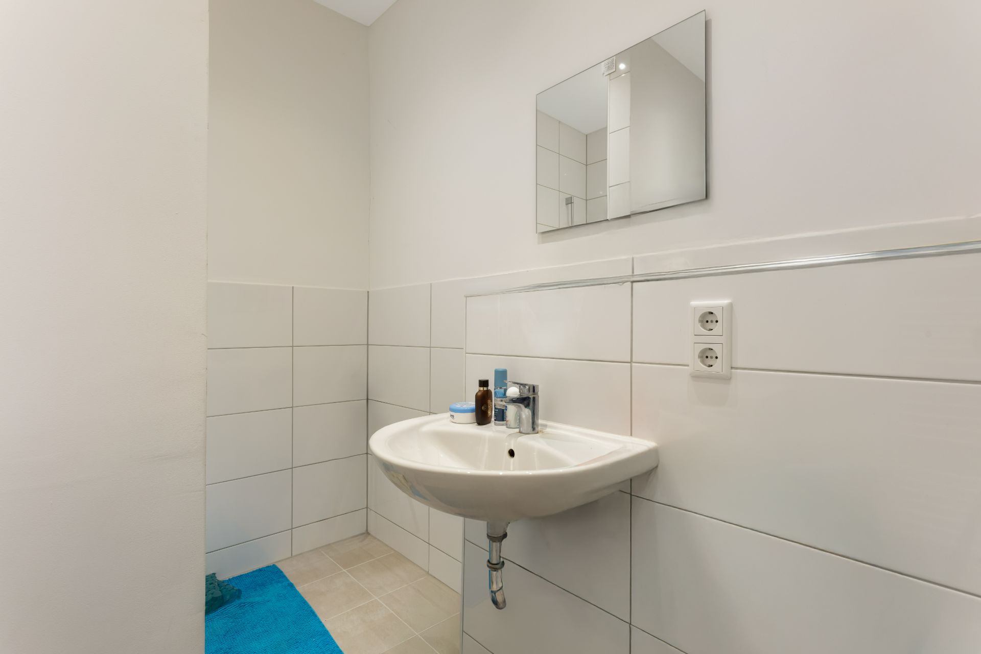 Helles Badezimmer mit Waschbecken, Spiegel und blauer Badematte auf Fliesenboden.