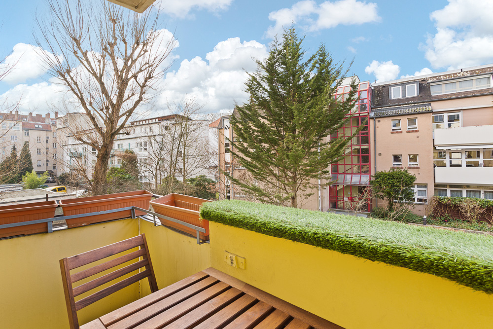 Kleiner Balkon mit Holztisch und Stuhl, Blick auf Bäume und umliegende Wohngebäude.