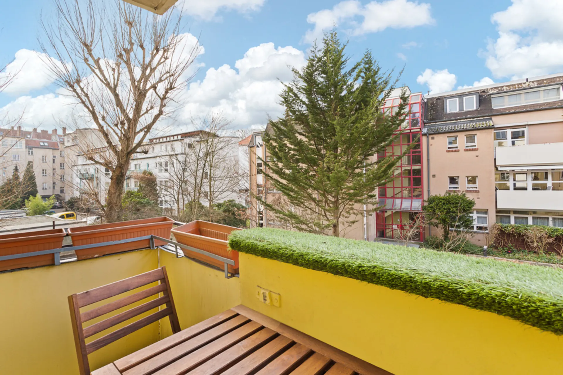 Kleiner Balkon mit Holztisch und Stuhl, Blick auf Bäume und umliegende Wohngebäude.
