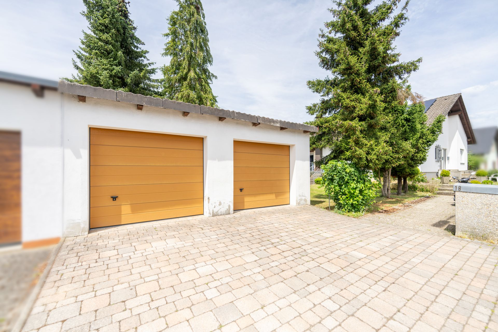 Doppelgarage mit gelben Toren und gepflasterter Einfahrt, daneben Bäume und Büsche.