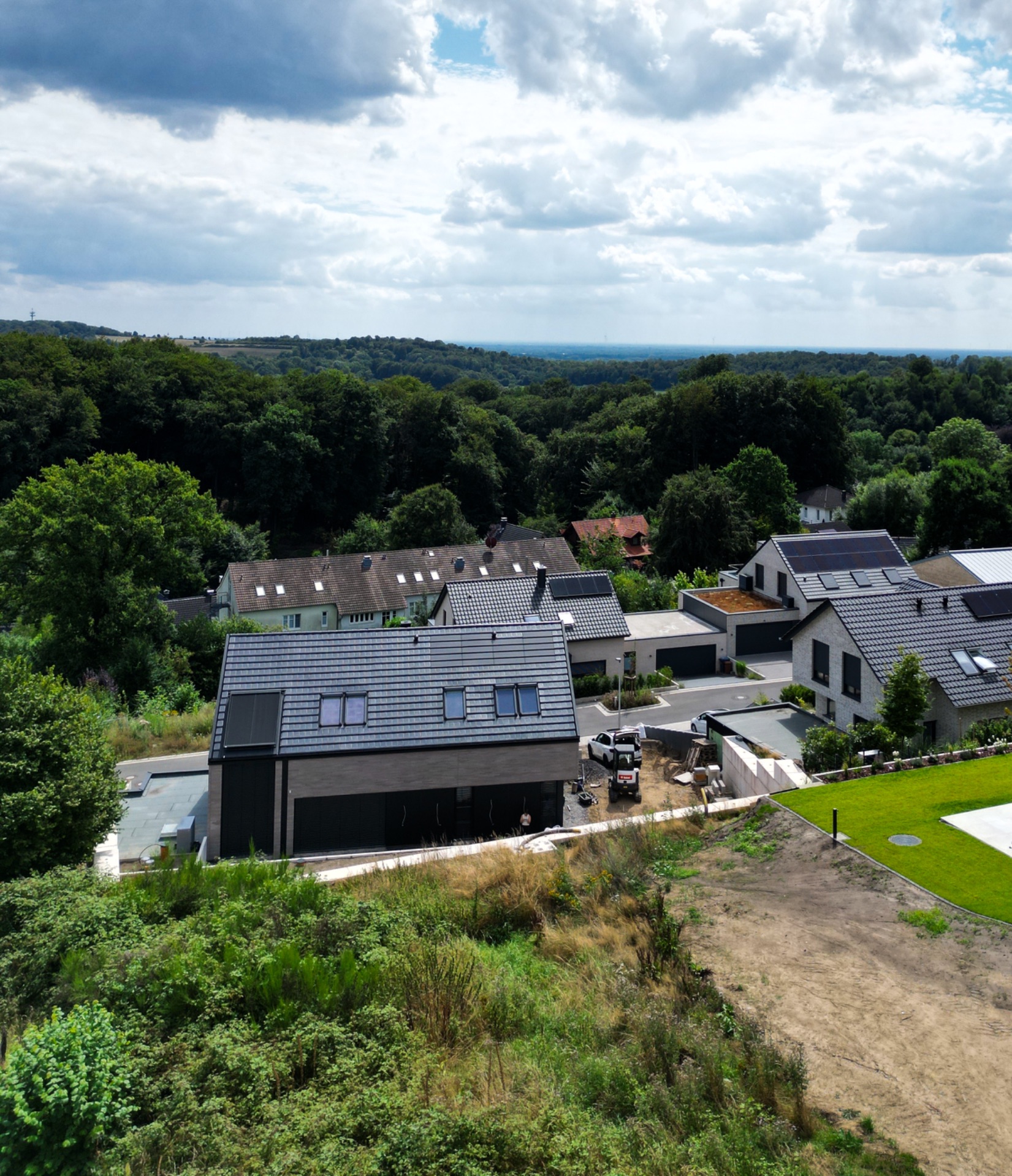 Moderne Wohnhäuser mit Gärten, umgeben von Bäumen und Wiese, unter teils bewölktem Himmel.