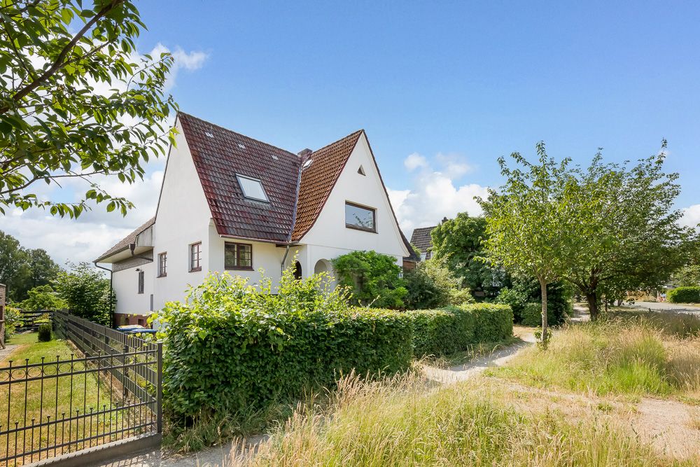 Weißes Einfamilienhaus mit rotem Dach, Garten und Bäumen an einer ruhigen Straße.