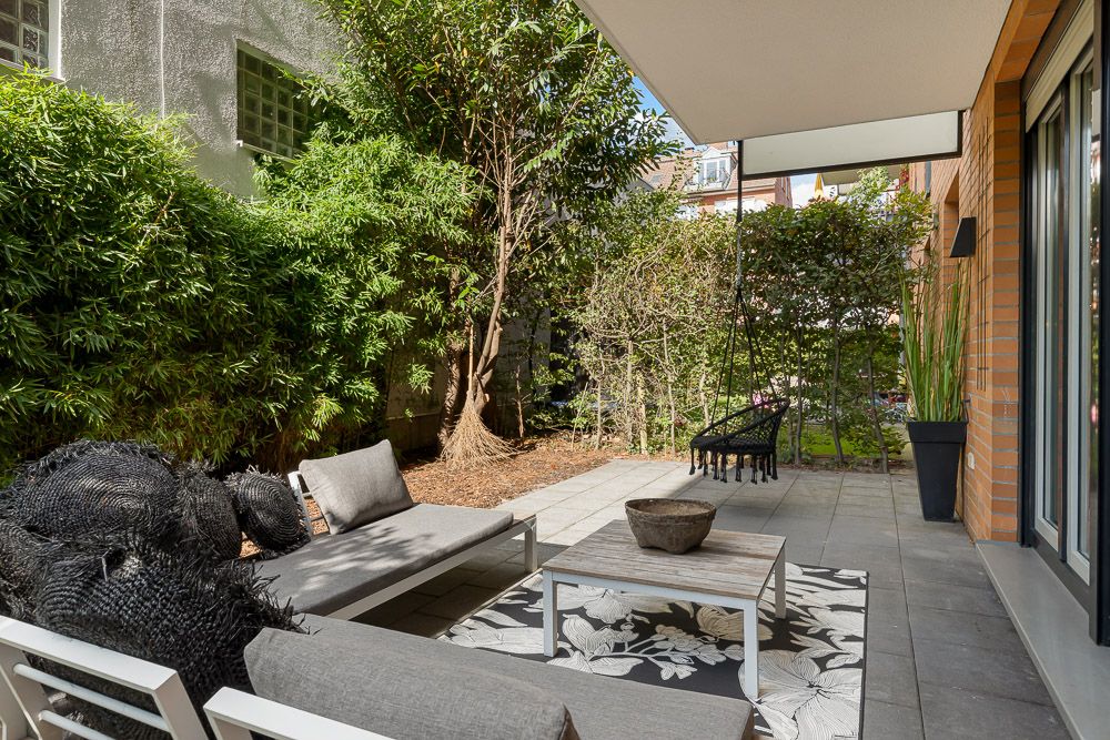 Moderne Terrasse mit grauen Gartenmöbeln, Teppich und viel Grünpflanzen am Rand.