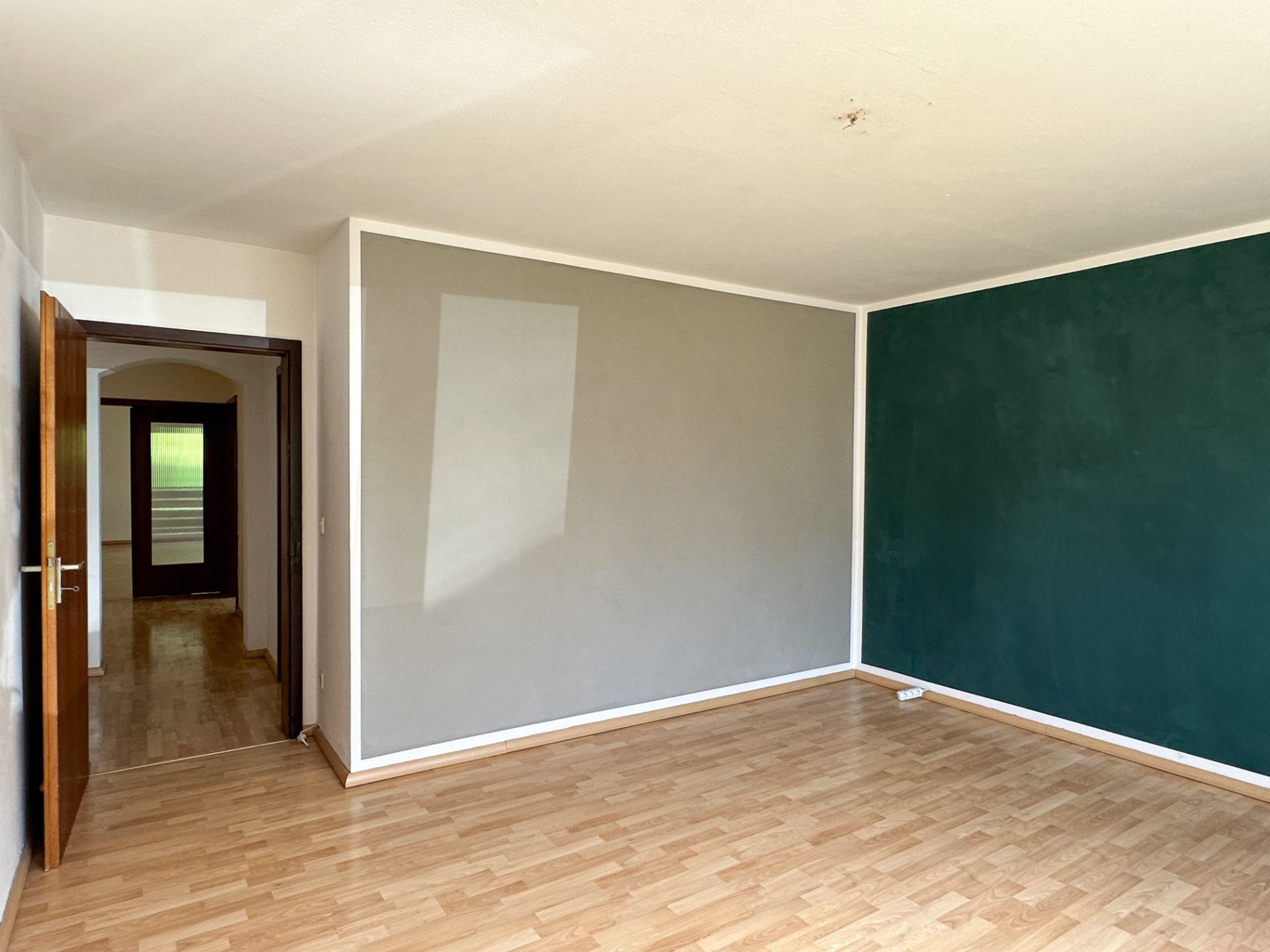 Leerer Raum mit Holzfußboden, grauer und grüner Wand, offene Tür zum Flur.