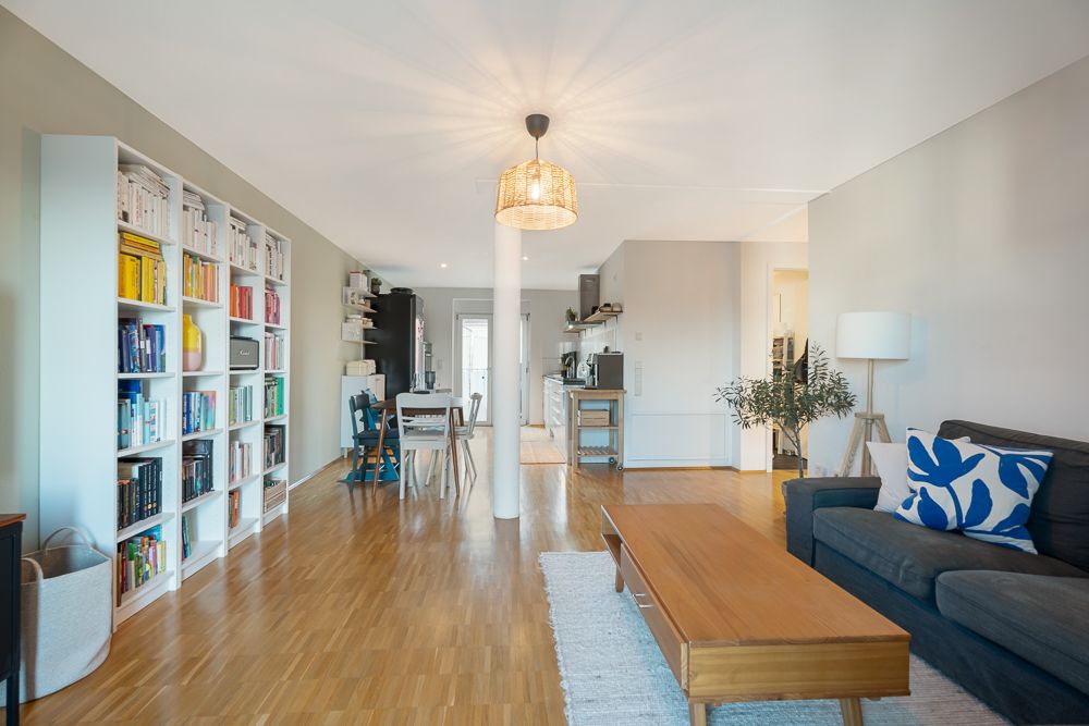 Helles Wohnzimmer mit Sofa, Couchtisch, Bücherregal und offener Küche im Hintergrund.