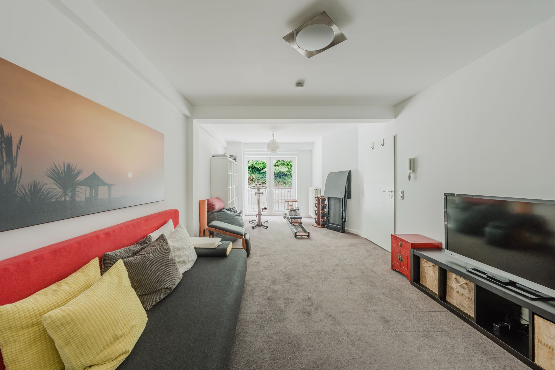 Helles Wohnzimmer mit Sofa, Fernseher und Zugang zu einem Balkon mit Blick auf Grünfläche.