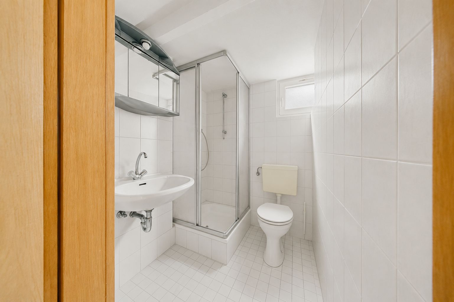 Helles Badezimmer mit Dusche, WC, Waschbecken und kleinem Fenster, komplett weiß gefliest.