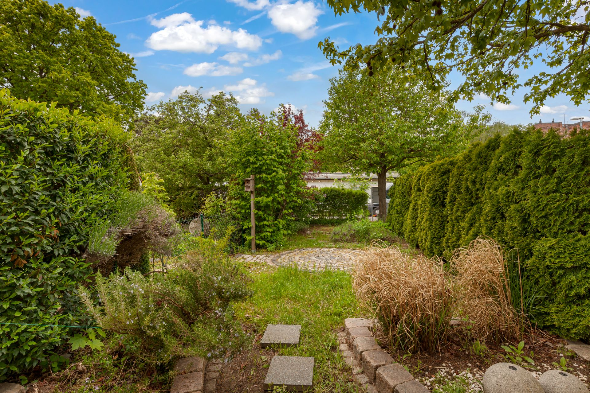 Garten mit Gehwegplatten, Sträuchern, Bäumen und einer kleinen gepflasterten Fläche im Hintergrund.