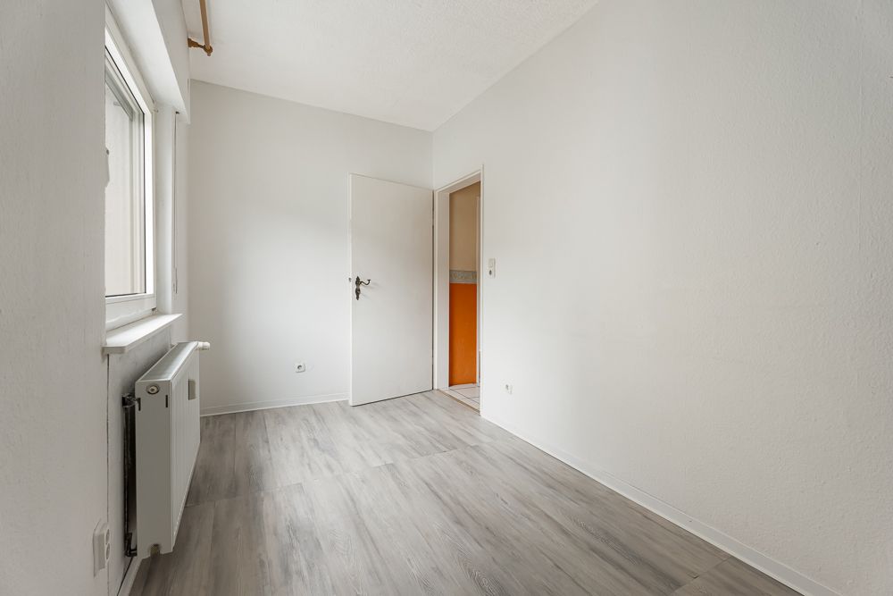 Helles, leeres Zimmer mit Fenster, Heizung und offener Tür zu einem orangefarbenen Raum.