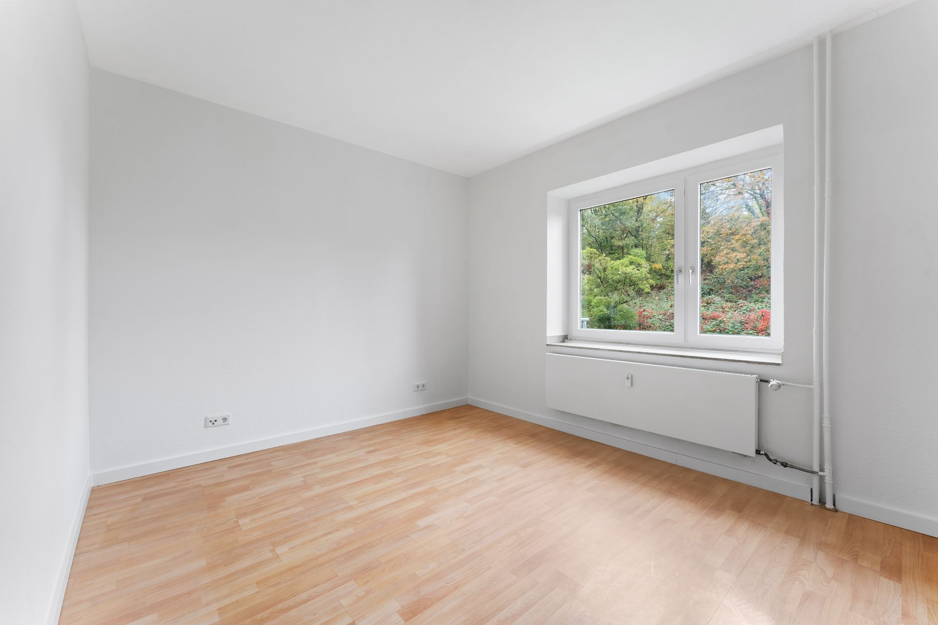 Helles, leeres Zimmer mit Holzfußboden und großem Fenster mit Blick ins Grüne.