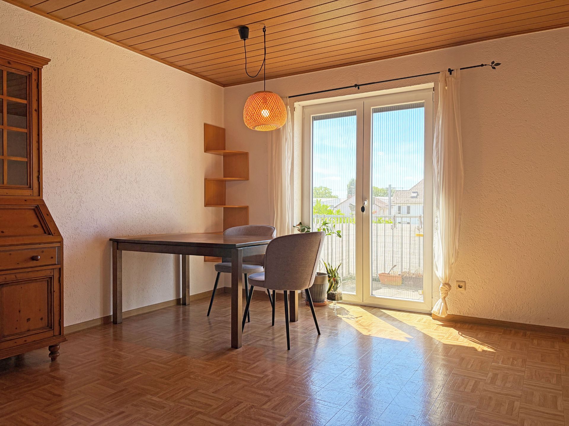 Helles Zimmer mit Holztisch, zwei Stühlen, Regal, Vitrine und Glastür zum Balkon.