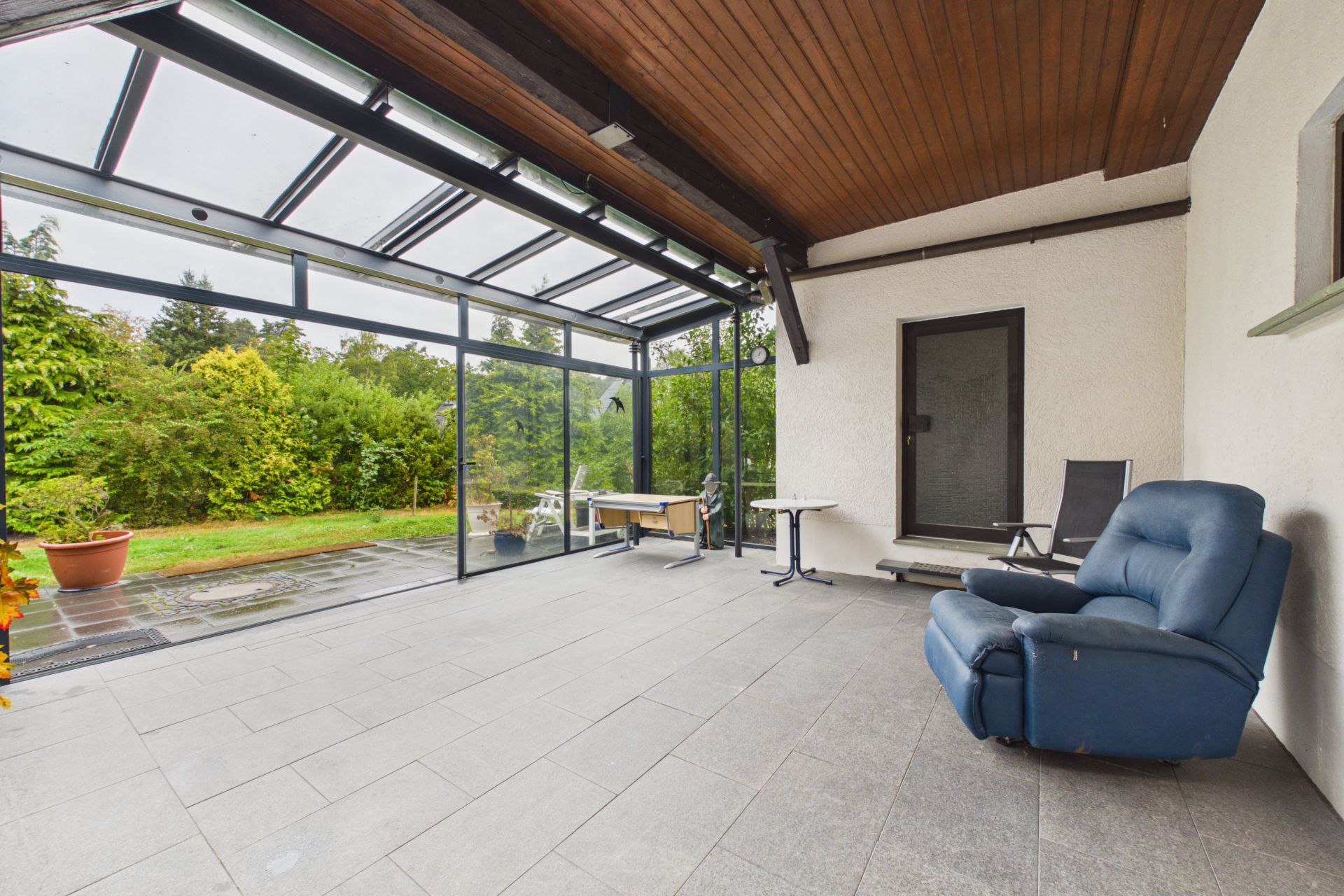 Überdachte Terrasse mit Glaswänden, Gartenblick, blauem Sessel und kleinen Tischen.