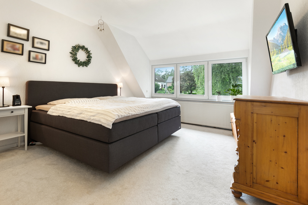 Helles Schlafzimmer mit großem Bett, Holzkommode, Fenstern und Wandfernseher.